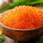 A close-up view of vibrant orange salmon roe in a wooden bowl, accompanied by lemon slices and fresh dill on a wooden surface.