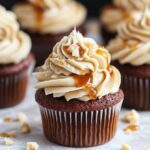 A close-up of a chocolate cupcake topped with salted caramel frosting, caramel drizzle, and crumbs, surrounded by similar cupcakes in the background.