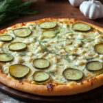 A gourmet white pizza topped with pickles, fresh dill, and chunks of garlic, served on a wooden board with garlic cloves and dill sprigs in the background.