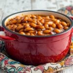 A red ceramic pot filled with baked beans in rich, glossy sauce, placed on a colorful patterned cloth over a rustic white wooden table.