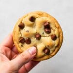 A close-up of a hand holding a freshly baked chocolate chip crumbl cookie with golden edges and melty chocolate chips.