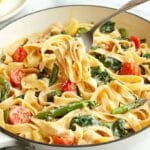 A skillet filled with creamy pappardelle pasta with chicken, asparagus, cherry tomatoes, and spinach, garnished with lemon slices.