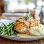 A plate of grilled chicken topped with creamy sauce, accompanied by green beans and mashed potatoes, served on a wooden table.